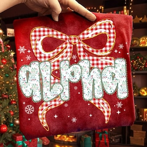 May include: A red t-shirt with a large bow design in red and white gingham. The word "ALPHA" is printed in a floral font with a green border. The design includes snowflakes and stars.