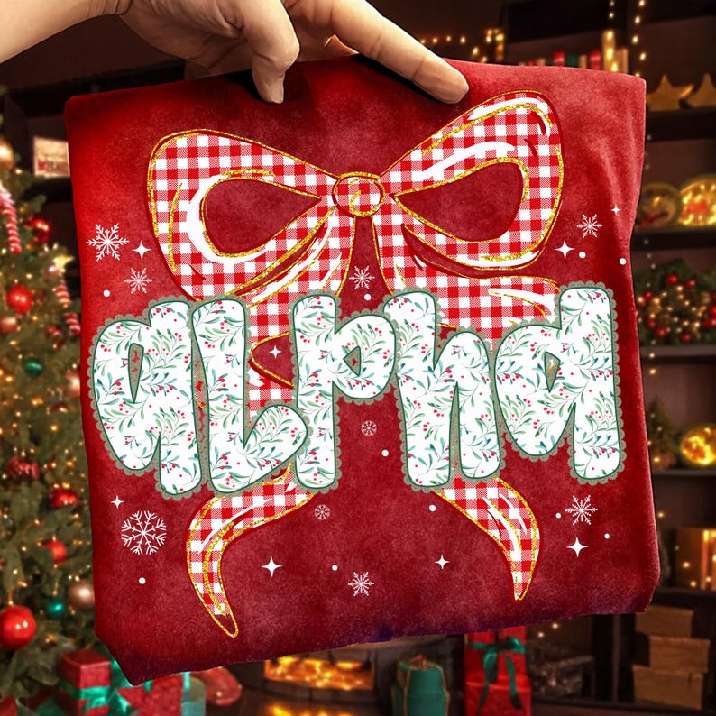 May include: A red t-shirt with a large bow design in red and white gingham. The word "ALPHA" is printed in a floral font with a green border. The design includes snowflakes and stars.