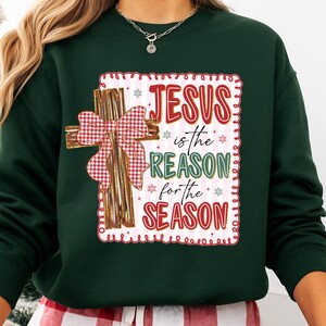 May include: Dark green sweatshirt with a decorative cross and the text "JESUS is the REASON for the SEASON" in red and green. The cross is brown with a red and white checkered bow. The text is inside a white frame with red trim.