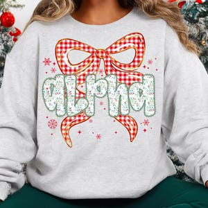 May include: A light gray sweatshirt featuring a large red and white plaid bow with gold trim. The word "ALPHA" is printed in a whimsical font, decorated with green leaves and white flowers. Red snowflakes and accents complete the design.