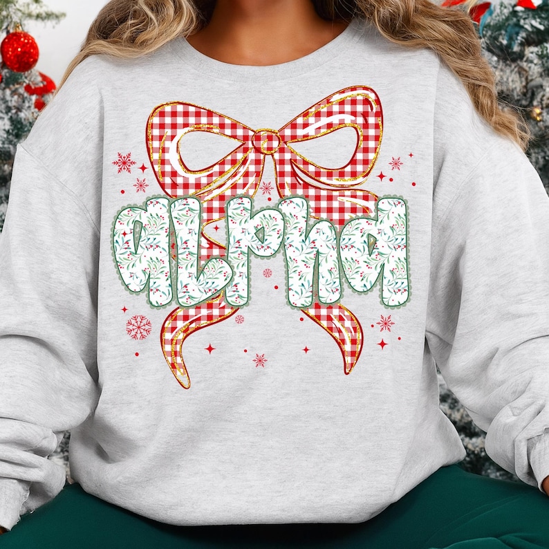 May include: A light gray sweatshirt featuring a large red and white plaid bow with gold trim. The word "ALPHA" is printed in a whimsical font, decorated with green leaves and white flowers. Red snowflakes and accents complete the design.