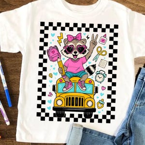 May include: White t-shirt featuring a cartoon raccoon wearing heart-shaped sunglasses and a pink bow, sitting on a yellow school bus. The raccoon is holding a pencil and making a peace sign. The design includes a pink backpack, scissors, and a clock.
