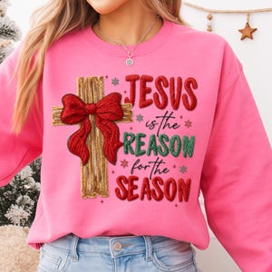May include: A pink sweatshirt with the text "JESUS is the REASON for the SEASON." The design includes a wooden cross with a red bow. The sweatshirt is a crew neck style.