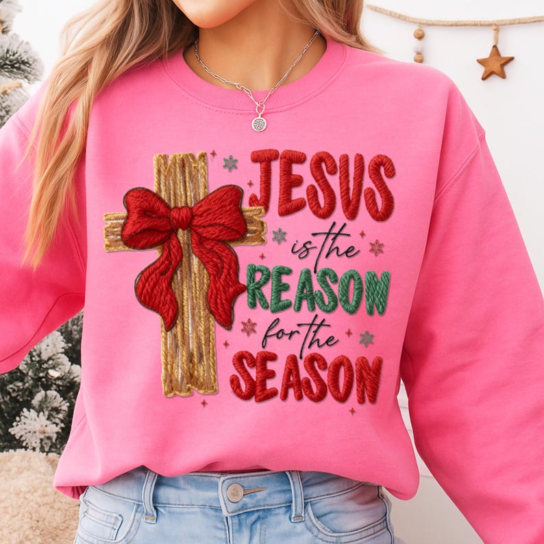 May include: A pink sweatshirt with the text "JESUS is the REASON for the SEASON." The design includes a wooden cross with a red bow. The sweatshirt is a crew neck style.