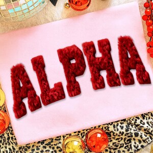 May include: A light pink textile with the word "ALPHA" in large, red, fuzzy letters. The letters are prominently displayed, with a soft, textured appearance. The background includes festive decorations and a leopard print.