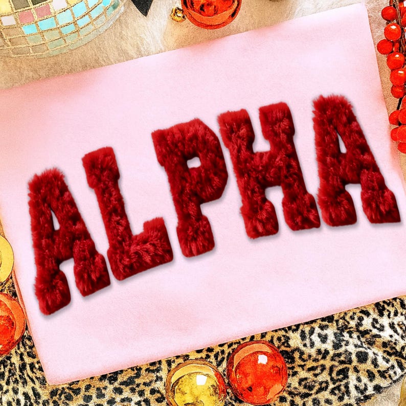 May include: A light pink textile with the word "ALPHA" in large, red, fuzzy letters. The letters are prominently displayed, with a soft, textured appearance. The background includes festive decorations and a leopard print.