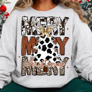 May include: A light gray sweatshirt with a Christmas tree design. The tree is white with black spots, antlers, and a gold bow. The words "MERRY CHRISTMAS" are printed in brown and leopard print.
