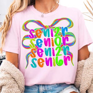 May include: A light pink t-shirt with the word "SENIOR" in large, colorful letters. The word is repeated four times, each in a different color, with a rainbow-colored bow design above. The shirt is a casual style.