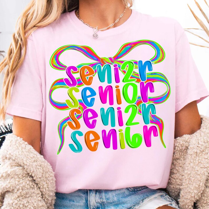 May include: A light pink t-shirt with the word "SENIOR" in large, colorful letters. The word is repeated four times, each in a different color, with a rainbow-colored bow design above. The shirt is a casual style.