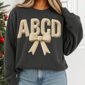 May include: Dark gray sweatshirt featuring the letters ABCD in a beige, knitted style, with a matching bow beneath. The sweatshirt has long sleeves and a crew neck. The design is centered on the front of the garment.