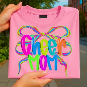 May include: A pink sweatshirt with the words "Cheer Mom" in colorful bubble letters. A rainbow-colored bow design is above the text. The sweatshirt is folded and held by hands.
