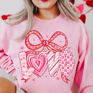 May include: A pale pink sweatshirt featuring a large red bow and the word "LOVE" in a heart-shaped design. The letters are decorated with stripes, hearts, and a checkered pattern. Small pink hearts are scattered around the design.