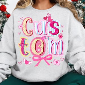 May include: A light gray sweatshirt with the word "CUSTOM" in a colorful, whimsical design. The letters are decorated with hearts, bows, and other Valentine's Day themed elements. The sweatshirt is a comfortable, casual style.