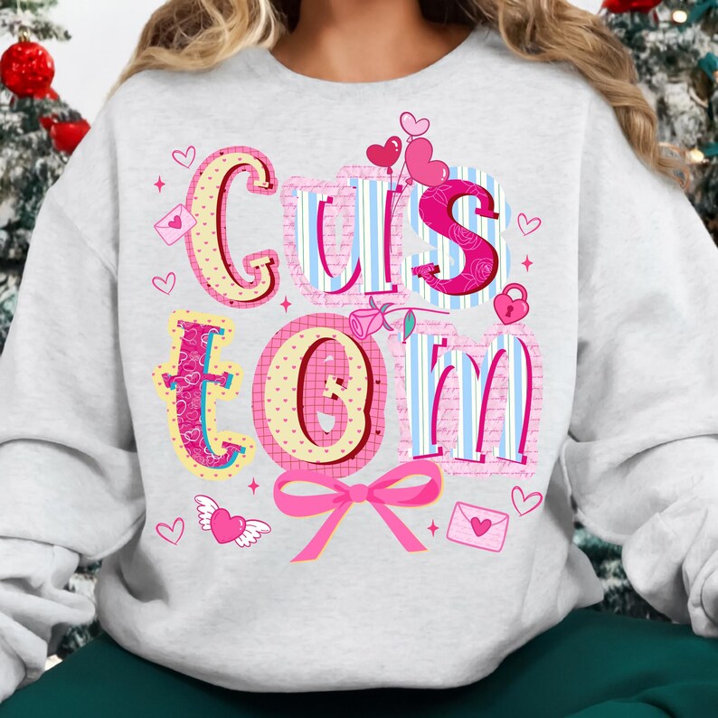 May include: A light gray sweatshirt with the word "CUSTOM" in a colorful, whimsical design. The letters are decorated with hearts, bows, and other Valentine's Day themed elements. The sweatshirt is a comfortable, casual style.