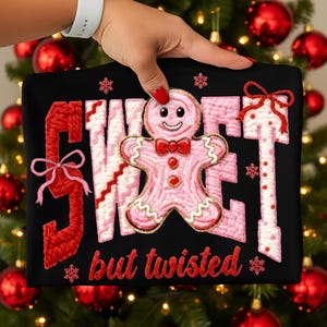 May include: Black sweatshirt with the words "SWEET" and "TWISTED" in red and pink yarn-like lettering. A pink gingerbread man with a red bow tie is in the center. Snowflakes and bows are also featured in the design.