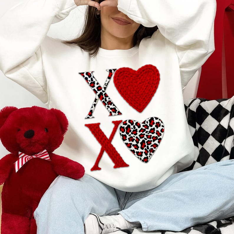 Może przedstawiać: Biała bluza z literami "XO", czerwonym sercem i sercem w panterkę. Po lewej stronie znajduje się czerwony pluszowy miś z muchą. Bluza ma czerwone i czarne "X" i serca w panterkę.