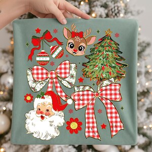 May include: A sage green cushion cover with Christmas illustrations. The design features a reindeer with a red bow, a decorated Christmas tree, Santa Claus, red and white checkered bows, baubles, and snowflakes.