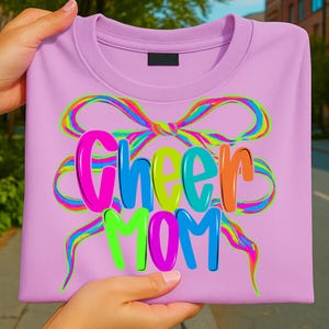 May include: A light purple sweatshirt with the words "Cheer Mom" in colorful bubble letters. A large, rainbow-colored bow design surrounds the text. The sweatshirt is made of a soft material.