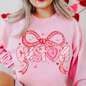May include: A light pink sweatshirt with a large red bow and the word "CUSTOM" in a decorative font. The letters are filled with patterns and a heart-shaped design. Small red hearts are scattered around the design.