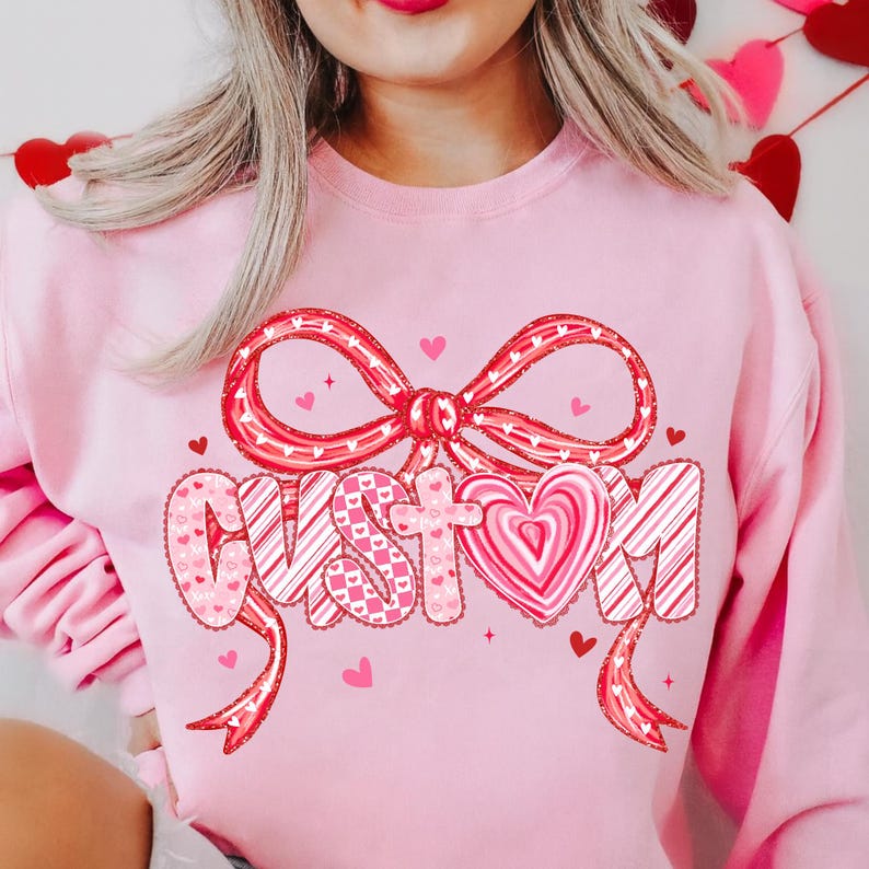 May include: A light pink sweatshirt with a large red bow and the word "CUSTOM" in a decorative font. The letters are filled with patterns and a heart-shaped design. Small red hearts are scattered around the design.