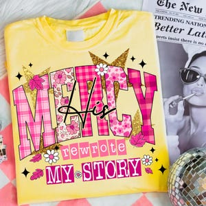 Op de afbeelding: Geel t-shirt met de tekst "MERCY His rewrote MY STORY" in roze, bloemen en gouden letters. Het ontwerp bevat bloemen- en ruitpatronen, met kleine bloemen- en bladaccenten. Het shirt ligt op een roze en wit gestreept oppervlak.