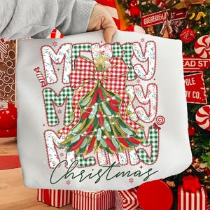 May include: White tote bag with a Christmas tree design. The words "MERRY CHRISTMAS" are printed in a festive font, with a red and green plaid bow and tree. The tree is decorated with red and gold accents.