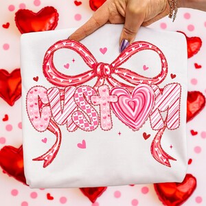 May include: White t-shirt with a large, decorative bow and the word "CUSTOM" in pink and red lettering. The letters have heart and candy cane stripe patterns. Small hearts and stars are scattered around the design.