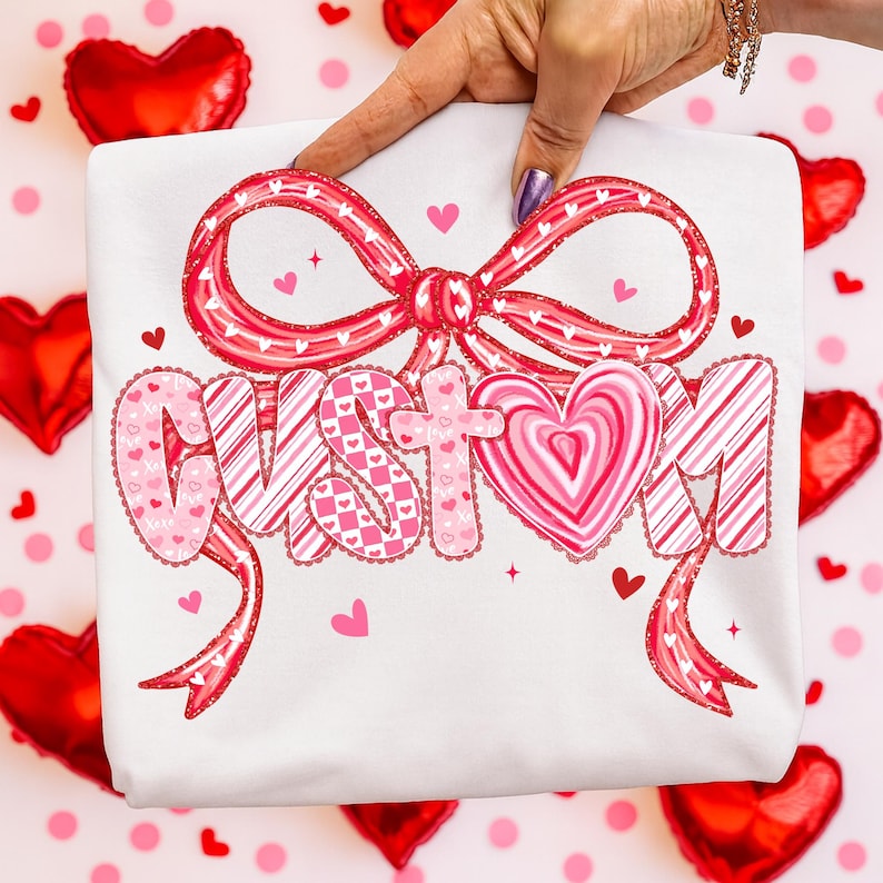 May include: White t-shirt with a large, decorative bow and the word "CUSTOM" in pink and red lettering. The letters have heart and candy cane stripe patterns. Small hearts and stars are scattered around the design.