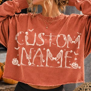 May include: A rust-colored long-sleeve crewneck sweatshirt with a raw-edge hem. The front features white graphic text that reads CUSTOM NAME, with letters formed by skeleton figures, surrounded by small bat, star, and jack-o'-lantern illustrations.
