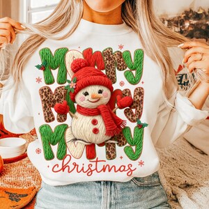 May include: White sweatshirt with a festive design featuring a snowman wearing a red hat and scarf. The words "Merry Christmas" are displayed in green, leopard print, and red lettering, with snowflakes.