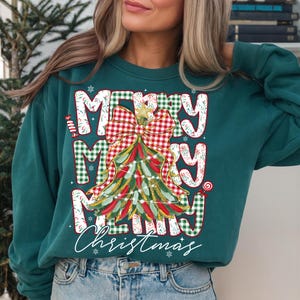 May include: Teal green sweatshirt with a festive Christmas tree design. The words "MERRY MERRY MERRY Christmas" are printed on the front in a mix of plaid and white lettering. The tree is decorated with lights and a bow.