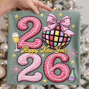 May include: Embroidered New Year's design on a sage green background. The design features the numbers "2026" in pink, a disco ball, champagne glass, fireworks, stars, and the words "Happy New Year" in gold.
