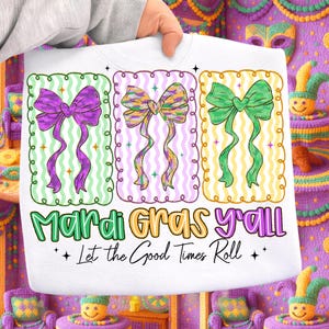 May include: White sweatshirt featuring three colorful bow designs in green, purple, and yellow, each within a decorative frame. The text "Mardi Gras y'all" and "Let the Good Times Roll" are printed below the bows, celebrating the festive season.