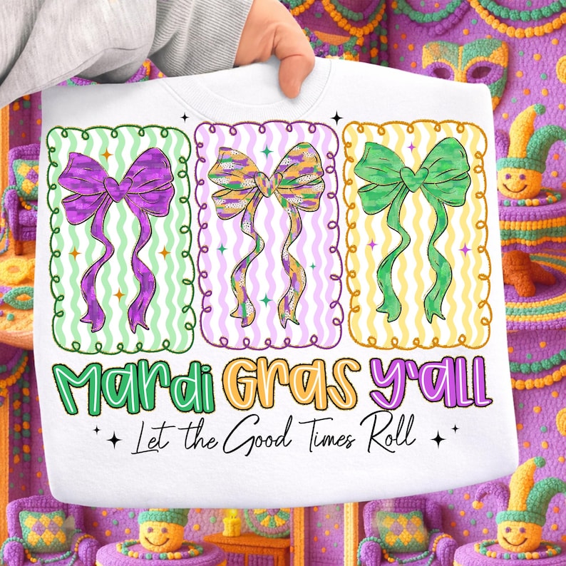 May include: White sweatshirt featuring three colorful bow designs in green, purple, and yellow, each within a decorative frame. The text "Mardi Gras y'all" and "Let the Good Times Roll" are printed below the bows, celebrating the festive season.