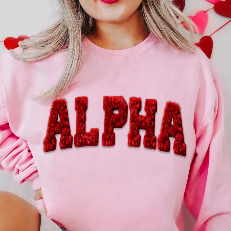 May include: A light pink sweatshirt with the word "ALPHA" in large, red, fuzzy letters across the front. The sweatshirt has a crew neck and long sleeves. The background includes decorative hearts.