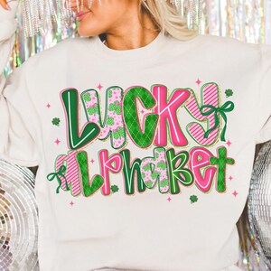 Pu&ograve; includere: Felpa color crema con le parole "LUCKY ALPHABET" in un design colorato. Il testo presenta lettere verdi e rosa con motivi a trifoglio e a righe, e fiocchi decorativi. Piccole stelle rosa e trifogli sono sparsi intorno al testo.