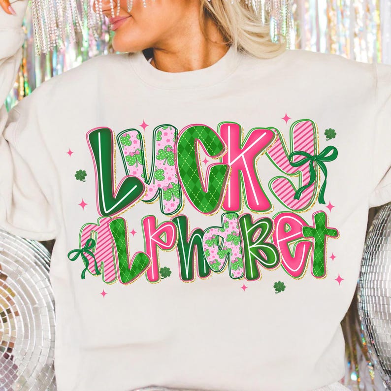 Pu&ograve; includere: Felpa color crema con le parole "LUCKY ALPHABET" in un design colorato. Il testo presenta lettere verdi e rosa con motivi a trifoglio e a righe, e fiocchi decorativi. Piccole stelle rosa e trifogli sono sparsi intorno al testo.