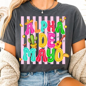 May include: Dark gray t-shirt with the text "ALPHA DE MAYO" in colorful letters. The design includes a sombrero, cactus, guitar, maracas, and other festive elements, set against pink and white stripes.