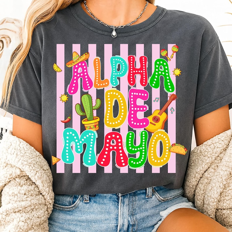 May include: Dark gray t-shirt with the text "ALPHA DE MAYO" in colorful letters. The design includes a sombrero, cactus, guitar, maracas, and other festive elements, set against pink and white stripes.
