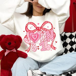 May include: White sweatshirt with a Valentine's Day graphic. The graphic features a large red bow, a pink heart, and the words "JOIN 3 16" in pink and white. A red teddy bear with a bow tie is on the left.