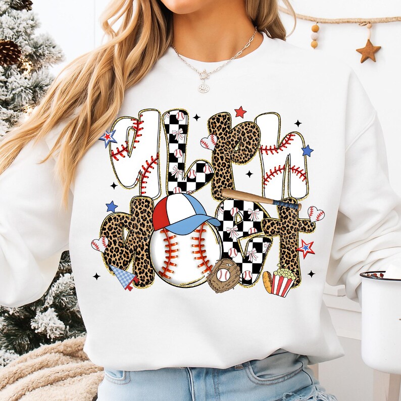 May include: White sweatshirt with a baseball-themed graphic. The design includes the words "Mom" and "Softball" in various fonts, baseballs, a baseball cap, and a leopard print pattern. The graphic also features stars and a baseball bat.