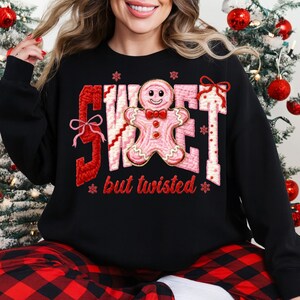 May include: Black sweatshirt with a festive design. The design features the word "SWEET" in red and pink, with a gingerbread man in the center. The text "but twisted" is below. Snowflakes and bows add to the holiday theme.