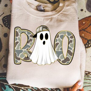 May include: A folded, light beige sweatshirt with a graphic that says "BOO". The letters are filled with a camouflage pattern and outlined in gold. The "O" is a ghost wearing a backwards baseball cap.