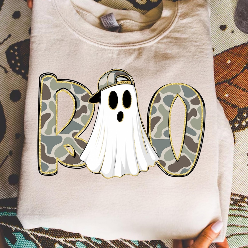 May include: A folded, light beige sweatshirt with a graphic that says "BOO". The letters are filled with a camouflage pattern and outlined in gold. The "O" is a ghost wearing a backwards baseball cap.