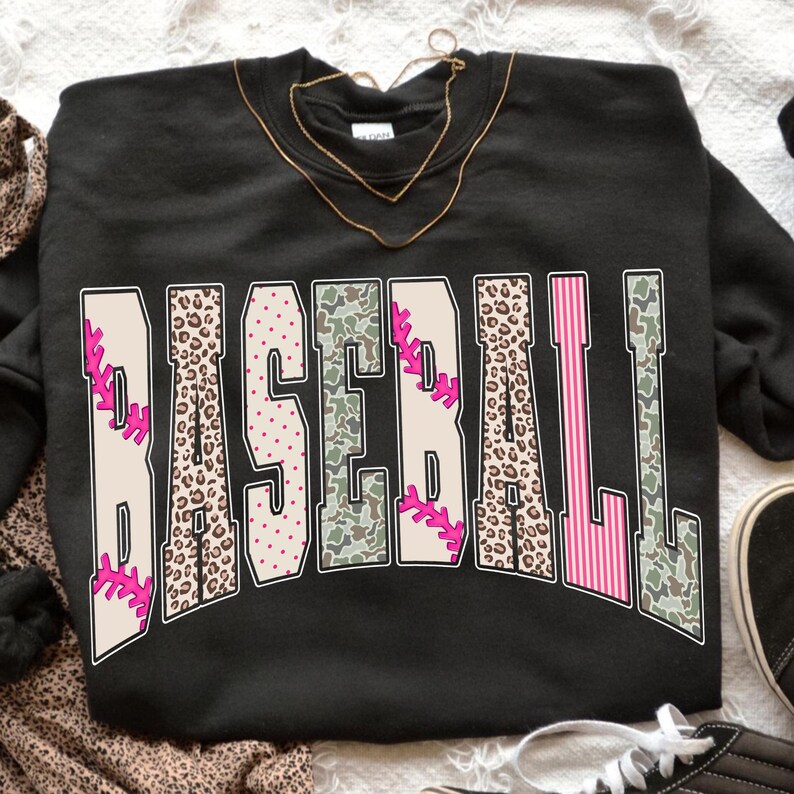 May include: Black sweatshirt with the word "BASEBALL" in a bold, arched design. The letters feature baseball-themed patterns, including pink stitching, leopard print, camouflage, and pink stripes. A gold necklace is draped on the sweatshirt.