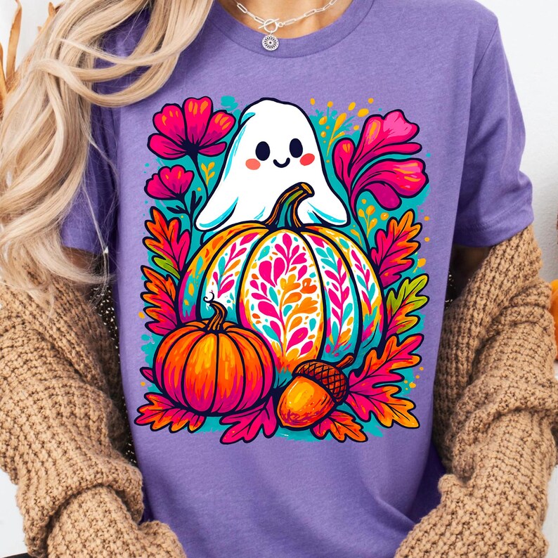 May include: Purple t-shirt featuring a colorful Halloween design. The graphic includes a white ghost, a large decorated pumpkin, a smaller orange pumpkin, and an acorn, surrounded by vibrant flowers and leaves.