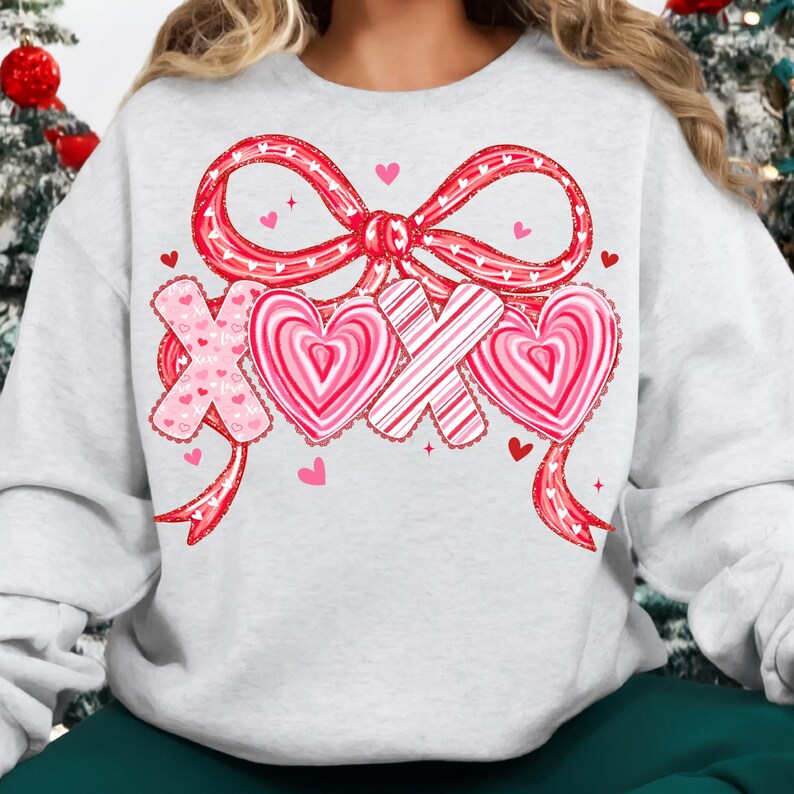 May include: A light grey sweatshirt with a red bow and the letters "XOX" in a Valentine's Day design. The "X" letters are pink with heart details, and the "O" letters are pink and white striped hearts.