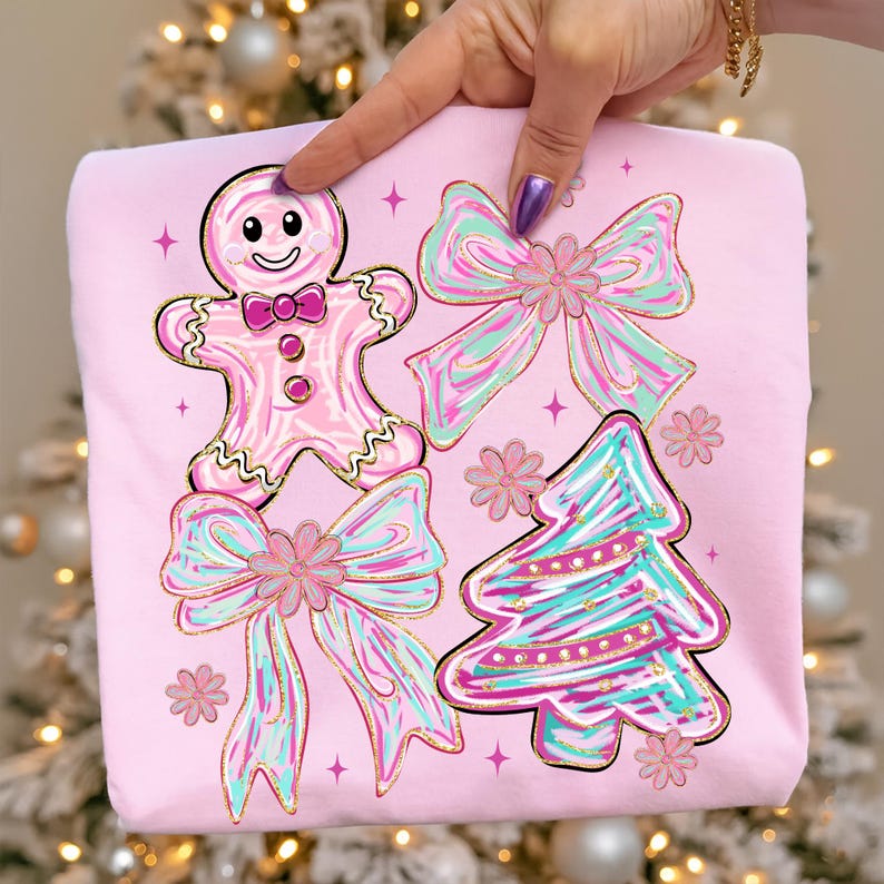 May include: A pale pink sweatshirt with a festive design. It features a gingerbread man, bows, and a Christmas tree, all with gold detailing. The bows and tree have teal and pink stripes. Pink stars and flowers are scattered around the design, creating a cheerful aesthetic.