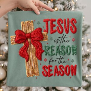 May include: A decorative pillow with the text "JESUS is the REASON for the SEASON." The pillow features a cross with a red bow and embroidered text in red and green on a teal background. The pillow is held by a person.