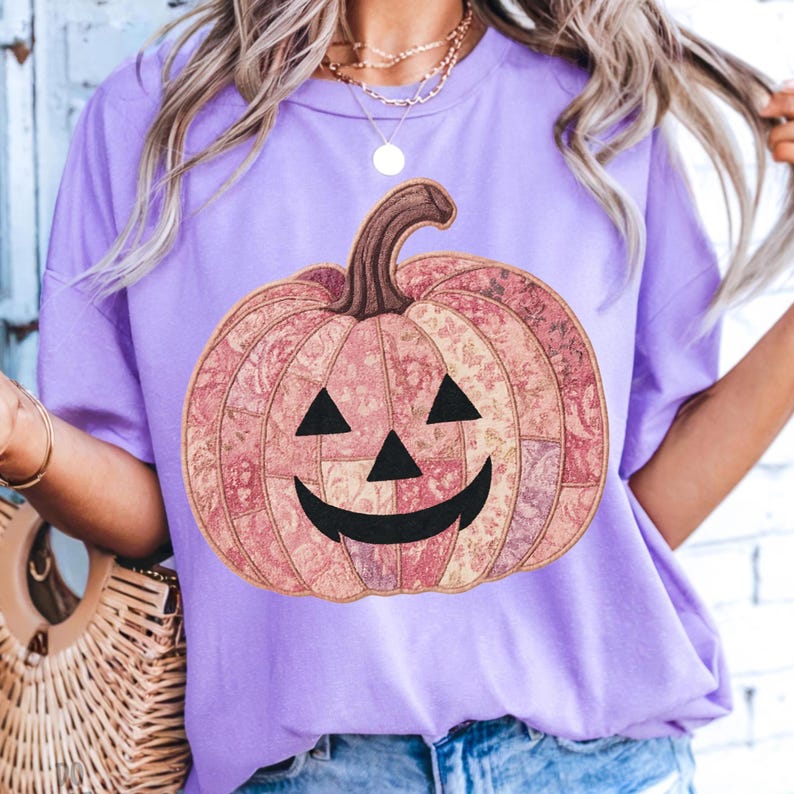 K&ouml;nnte beinhalten: Ein lila T-Shirt mit einem gro&szlig;en K&uuml;rbisdesign. Der K&uuml;rbis besteht aus Patchwork-Stoff in Rosa- und Beiget&ouml;nen, mit einem braunen Stiel, schwarzen Dreiecksaugen und einem l&auml;chelnden Mund. Ideal f&uuml;r Halloween.